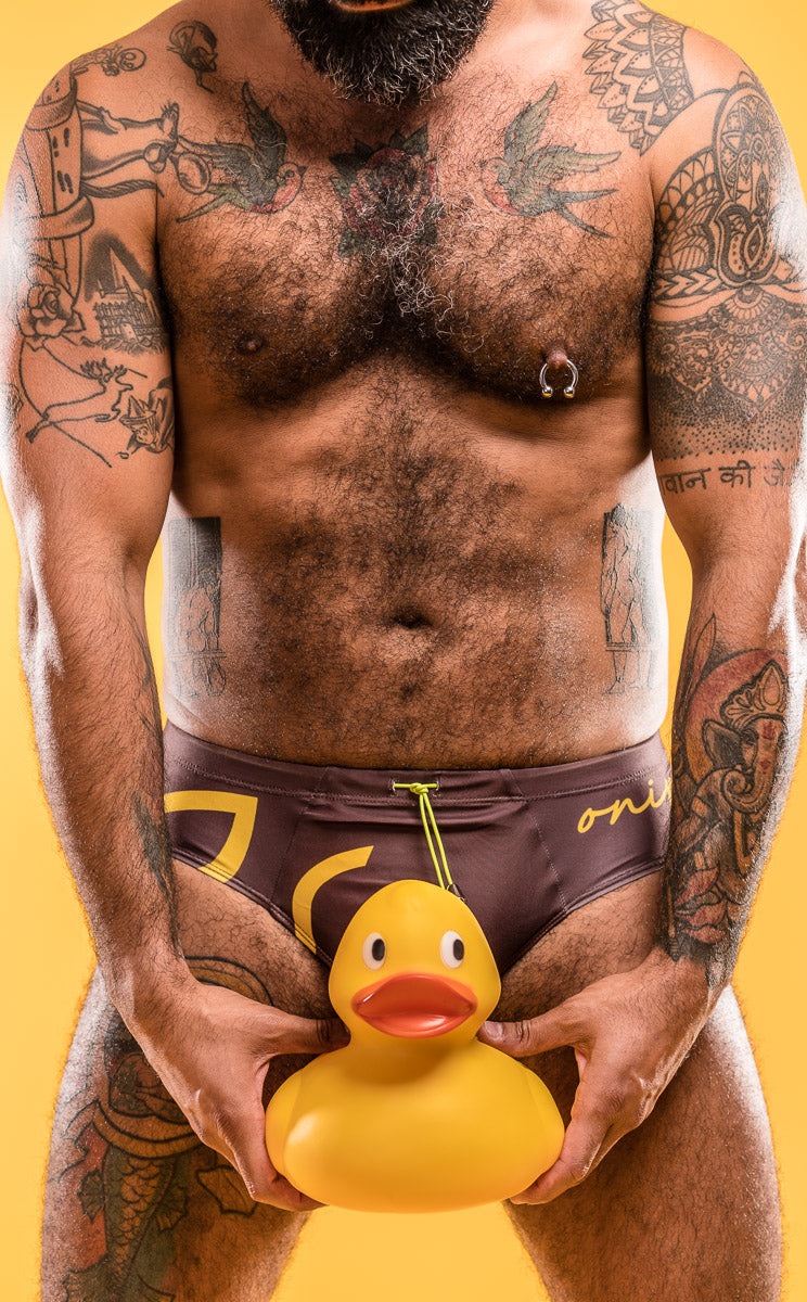 Man holding a yellow rubber duck in a brown swimwear with a yellow background